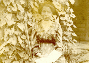 Historical image of an African American woman sitting on a chair next to lush vine growing next to a building